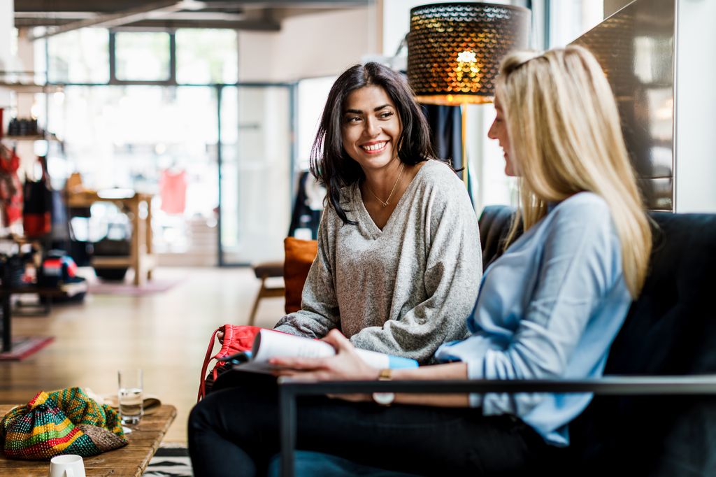 Two women go shopping during the day, taking a break and sitting on the couch in a clothing store together.