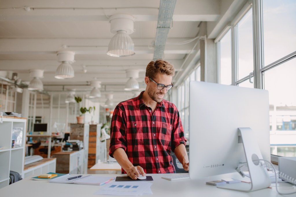 Image of a happy young man working on a desktop computer in a modern workplace.  Young entrepreneur working on start-up.