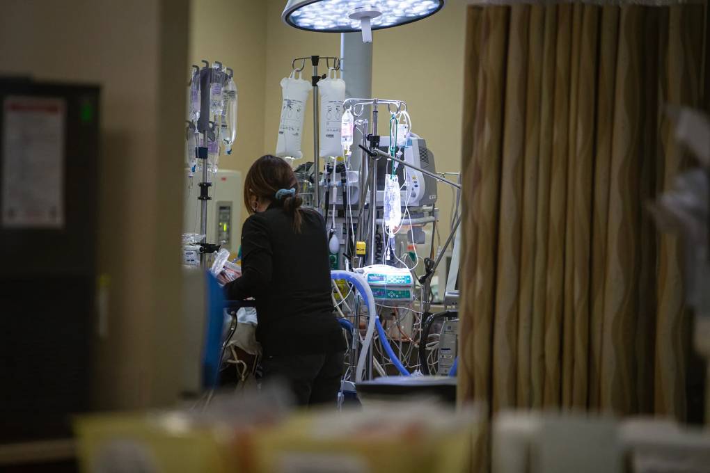 A female nurse is seen from behind, attending to a patient in a hospital room with medical equipment.