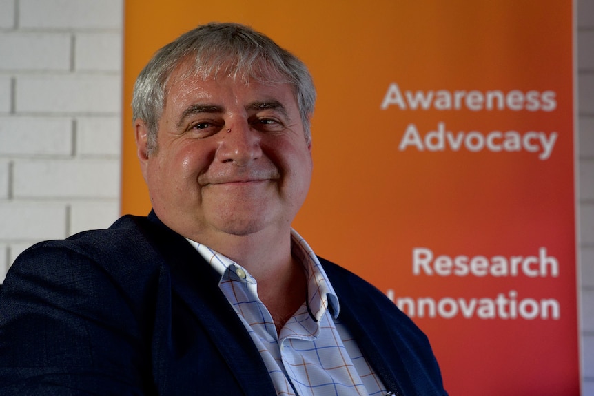 The man in the suit smiles in front of an orange banner with text that says advocacy for awareness and innovation in research.